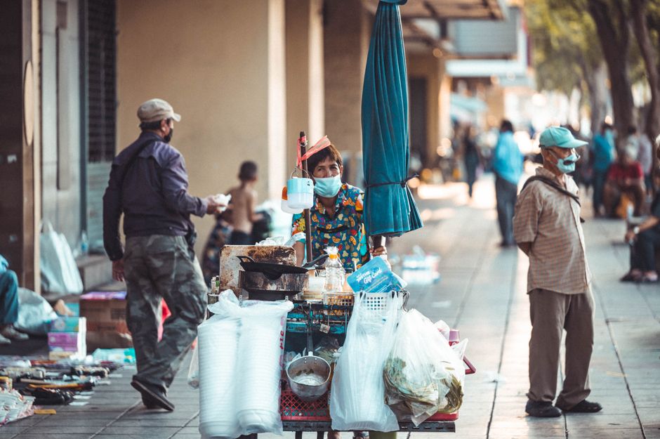 A bustling urban scene featuring a masked street vendor with a food cart and pedestrians.