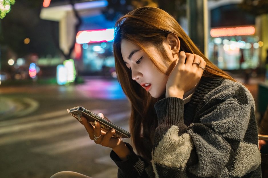 Asian woman with brown hair engrossed in her smartphone on urban city streets at night.