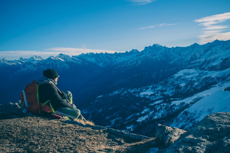 A man sits on a mountain ridge enjoying a breathtaking snow-capped mountain view under a clear blue sky.
