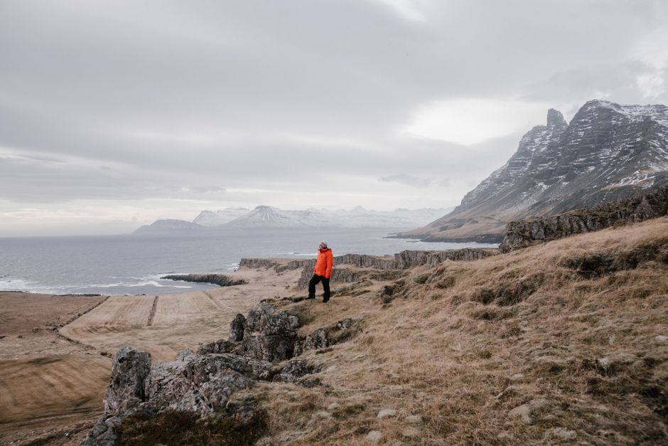 A man in a bright jacket hikes along a rugged sea cliff with snowy mountains in winter.