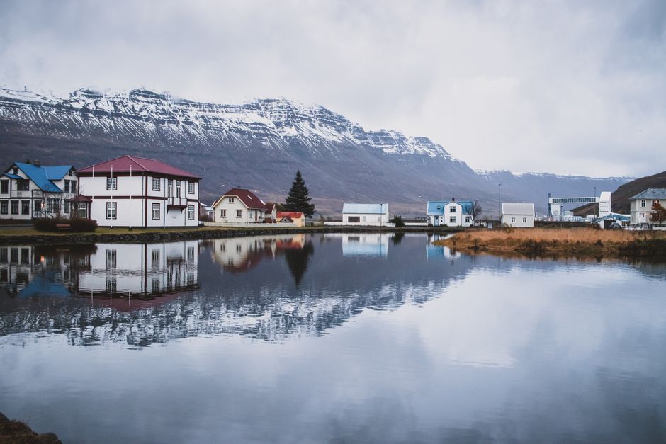 Picturesque Icelandic village with reflections on a calm lake, against a mountainous backdrop.