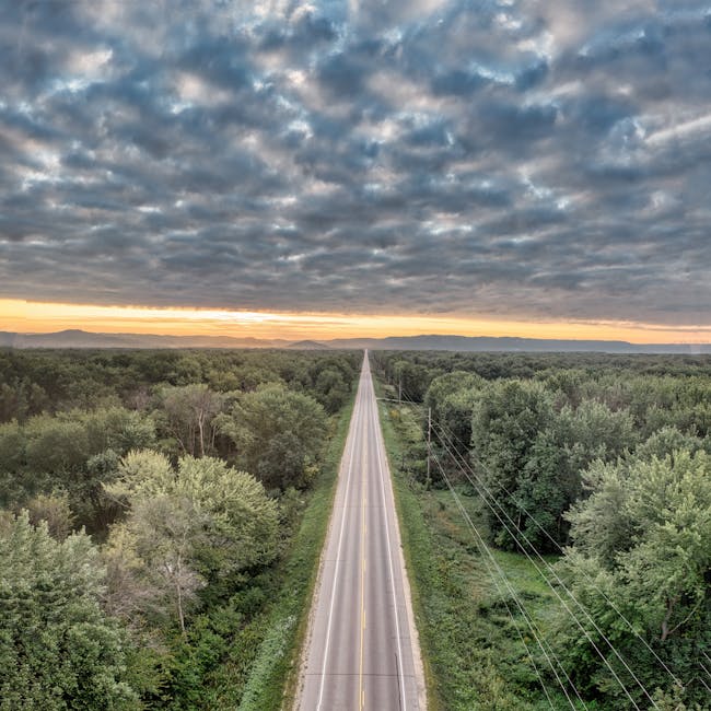 Discover a tranquil rural road through lush forests under a dramatic cloudy sky in Trevino, WI.