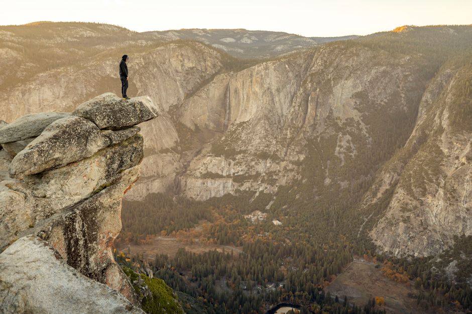 A lone adventurer stands on a cliff edge overlooking Yosemite Valley at sunset.