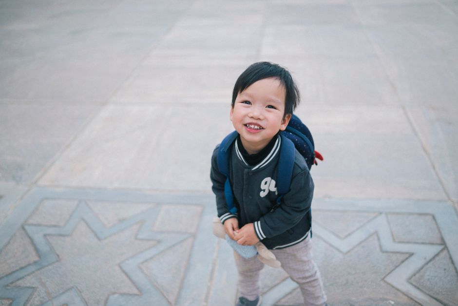 A cheerful child in a jacket and backpack smiles up at the camera on a concrete pavement.