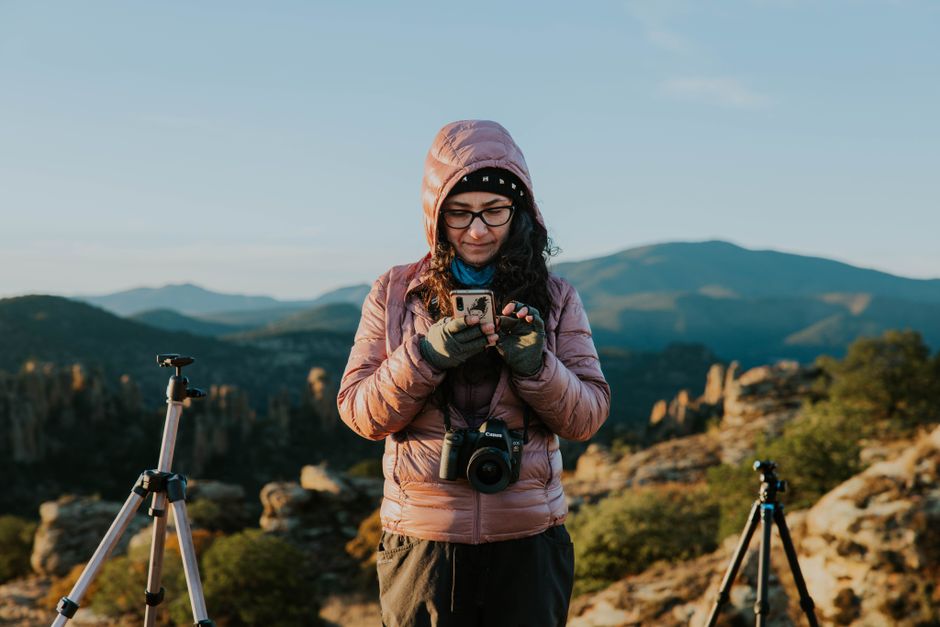 Female photographer in hooded jacket capturing stunning mountain scenery with a smartphone and tripod.