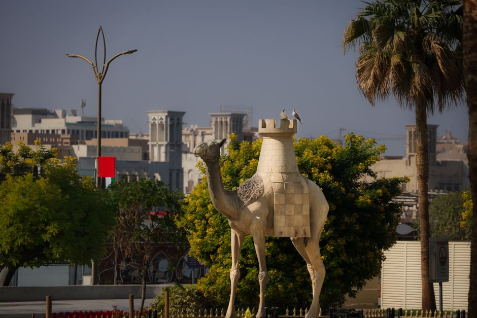 A camel statue surrounded by lush greenery and urban architecture under clear skies.